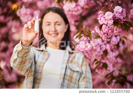 Focus on woman hand's holding nasal spray. Smiling woman allergic using medical nasal drops, suffering from seasonal allergy in blossoming garden on blurred background. Spring allergy concept. Focus on woman hand's holding nasal spray. Smiling woman allergic using medical nasal drops, suffering from seasonal allergy in blossoming garden on blurred background. Spring allergy concept. 122855780