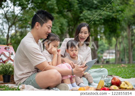 Asian parents are taking their kids on a picnic in a green park, watching a cartoon on a tablet. 122855854