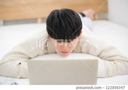 一名 20 多歲的年輕男子趴在床上玩電腦 122856347