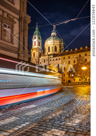 A tram moves swiftly in the heart of Prague's Lesser Town Square at night, illuminated by streetlights and the stunning architecture of baroque buildings in the backdrop. 122856645