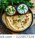 A rustic plate of savory pancakes garnished with fresh parsley, served with two small bowls of cream or yogurt.  122857328