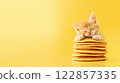 A sleepy kitten with soft orange fur gently rests its paws on a stack of pancakes, creating a peaceful and adorable scene. The yellow background adds to the warmth and calmness of the shot. 122857335