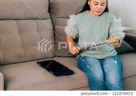 Female musician in a green top sits on a gray sofa at home, preparing her silver flute for a performance. The living room ambiance reflects her dedication and love for creating music. 122858953