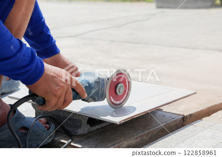 Precision and expertise: a worker in blue clothing uses an angle grinder to shape a gray tile on a construction site, showcasing careful attention to detail and craftsmanship. 122858961