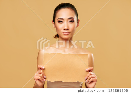 Young asian woman holding torn piece of blank paper against beige studio background. Young asian woman holding torn piece of blank paper against beige studio background. 122861596