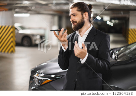 Young Caucasian male businessman recording audio message on smartphone while standing near car in parking garage. Wearing formal suit, projecting sense of professionalism and modern communication. Young Caucasian male businessman recording audio message on smartphone while standing near car in parking garage. Wearing formal suit, projecting sense of professionalism and modern communication. 122864188