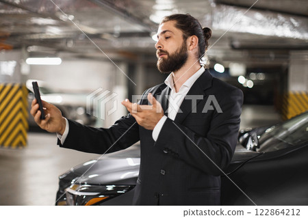 Young Caucasian male in suit speaking on phone video call in underground car park next to car. Business communication concept with focus on technology and modern work style. 122864212