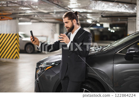 Young Caucasian male in suit speaking on phone video call in underground car park next to car. Business communication concept with focus on technology and modern work style. 122864224