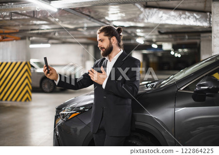 Caucasian male in 30s using smartphone for video call in underground parking lot, standing by car wearing black suit looks focused and professional, urban setting, modern communication business style Caucasian male in 30s using smartphone for video call in underground parking lot, standing by car wearing black suit looks focused and professional, urban setting, modern communication business style 122864225