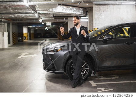 Young Caucasian male in suit uses smartphone for video call in garage, hot drink in hand, beside car. Young Caucasian male in suit uses smartphone for video call in garage, hot drink in hand, beside car. 122864229