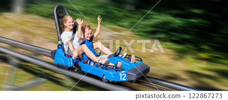 Pair of cute laughing kids enjoy have fun riding alpine roller coaster cart together at Harz nature park on sunny holidays summer day. Children summer outdoors travel active sports adventure 122867273