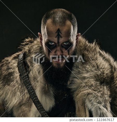 Close-up portrait of Viking warrior with war paint, piercing blue eyes, and fur cloak, staring menacingly against dark background. 122867617