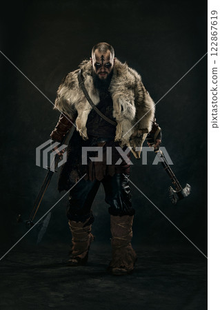 Fierce Viking warrior with battle scars and war paint gripping two axes, dressed in fur cloak and chainmail, staring intensely against dark background. 122867619