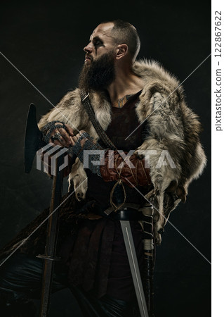 Portrait of man as medieval Viking, warrior dressed in fur cloak and armor holding battle axe while gazing into distance against dark background. 122867622