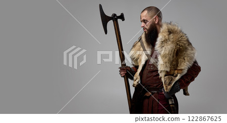 Medieval Viking warrior dressed in armor, with massive axe, showing fierce determination against grey studio background. 122867625