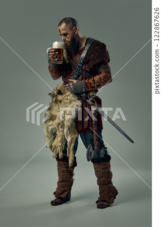 Man, Viking warrior dressed in fur cloak and leather armor, standing with sword, smelling beer, against studio background. 122867626