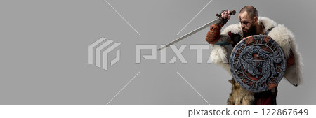 Moment of defense. Medieval warrior, Viking dressed in fur cloak and armor, standing with shield and sword against grey studio background 122867649