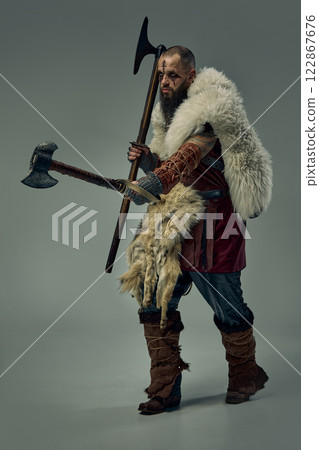 Strategy and determination. Full-length portrait of focused man, medieval warrior, Viking in full armor walking with two axes. 122867676