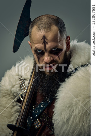 Portrait of man, Viking warrior smirking slightly, with battle axe, fierce expression and fur cloak against grey studio background. 122867681