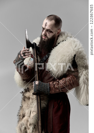 Brutal man, medieval Viking, warrior in full armor and fur cloak, examining battle axe against grey studio background. 122867685