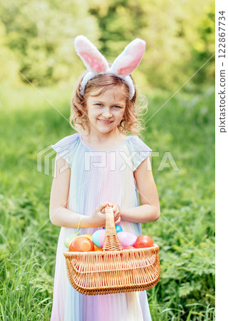 Easter egg hunt. Girl child Wearing Bunny Ears Running To Pick Up Egg In Garden. Easter tradition. Baby with basket full of colorful eggs. Bunny in basket Easter egg hunt. Girl child Wearing Bunny Ears Running To Pick Up Egg In Garden. Easter tradition. Baby with basket full of colorful eggs. Bunny in basket 122867734