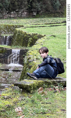 Hiker boy sits on green meadow near mountain stream Hiker boy sits on green meadow near mountain stream 122868895