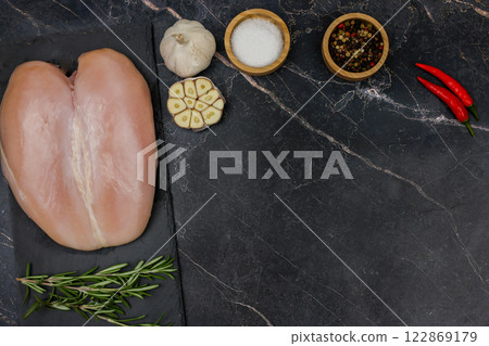 Chicken breast fillet lies on a black board on a dark background with a sprig of rosemary next to it, garlic, red pepper and spices and salt on top in wooden bowls with space for text. The concept of Chicken breast fillet lies on a black board on a dark background with a sprig of rosemary next to it, garlic, red pepper and spices and salt on top in wooden bowls with space for text. The concept of 122869179