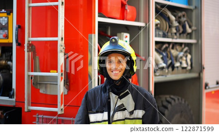 Portrait of happy female fireguard against the background of big red truck. Young firewoman in full equipment looking at camera with positive emotions near a fire engine. Concept of heroic profession 122871599