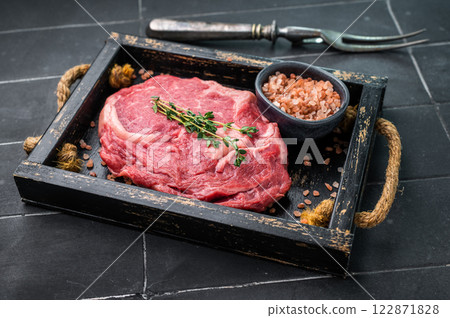 Marbled raw Rib eye steak with herbs in wooden tray, fresh beef meat. black kitchen background. top view Marbled raw Rib eye steak with herbs in wooden tray, fresh beef meat. black kitchen background. top view 122871828