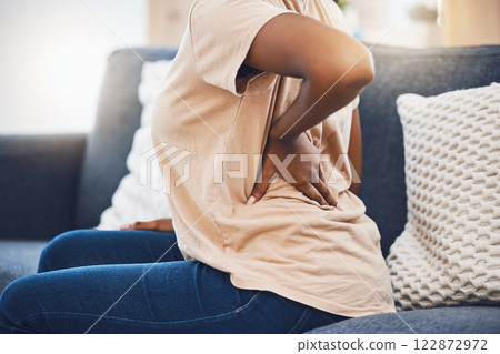 Woman with a back injury, pain or accident from stress sitting on sofa in her living room. Black girl with a muscle sprain, back pain or injured spine medical emergency on couch in lounge at home. 122872972