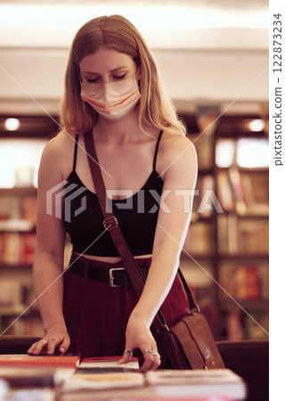 Covid, face mask and woman with books in a library, bookshop or educational building for knowledge, reading or research. Young female student browsing and choosing novel on table in a bookstore Covid, face mask and woman with books in a library, bookshop or educational building for knowledge, reading or research. Young female student browsing and choosing novel on table in a bookstore 122873234