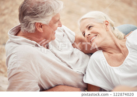 Senior couple, beach and vacation with a smile, love and together while relax on the sand together in summer. Elderly man and woman enjoy romantic holiday, anniversary or date at the ocean in nature 122874020