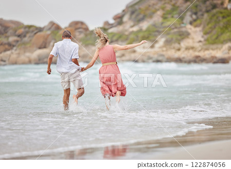 Interracial couple, ocean fun and holiday vacation together holding hands and enjoying the water. Man and woman bonding in a joyful relationship for beach time, waves and nature in the outdoors 122874056