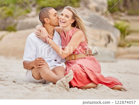 Beach, multiracial love and a couple kiss in sand while sitting on ocean holiday or honeymoon. A black man and happy woman with smile enjoying nature, weekend and summer vacation at the sea together. 122874057