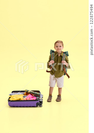 Boy standing with backpack on his shoulders, holding straps, suitcase with clothes and map beside him, against yellow background. 122875494