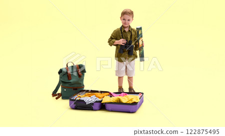 Boy standing, holding map and binoculars, suitcase with clothes nearby, green backpack beside him, against yellow background. 122875495