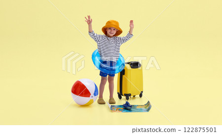 Boy joyfully raising hands, wearing inflatable ring, suitcase nearby, beach ball, map, glasses, and toy ship around against yellow background. 122875501