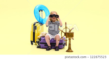Exploration. Girl sitting on suitcase, looking through binoculars near travel bags, inflatable ring and ship wheel Exploration. Girl sitting on suitcase, looking through binoculars near travel bags, inflatable ring and ship wheel 122875523