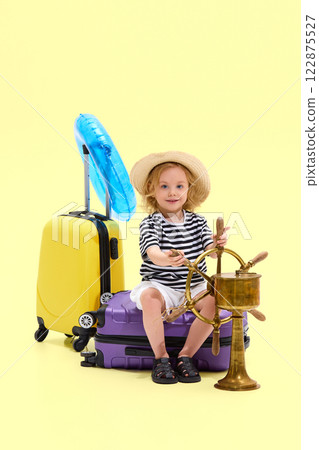 Girl sitting on suitcase in striped outfit and straw hat, gripping ship wheel, pretending to captain voyage Girl sitting on suitcase in striped outfit and straw hat, gripping ship wheel, pretending to captain voyage 122875527
