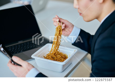 A young man in his twenties eating cup noodles while operating a smartphone 122875587