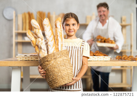 In working room of bakery, there is apprentice girl with baguettes in wicker basket in hands In working room of bakery, there is apprentice girl with baguettes in wicker basket in hands 122875721
