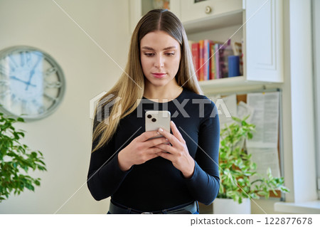 Young smiling woman using smartphone standing at home 122877678
