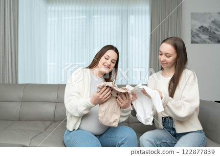 Young pregnant woman and her female friend choosing baby clothes while sitting on a couch. Concept of motherhood, friendship, preparation, excitement, and bonding. 122877839