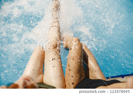 Cropped view of two pair of long tanned female legs sitting near blue sparkling water of the pool. Girls wearing black and khaki swimwear. Female friends chilling near swimming pool. 122878045