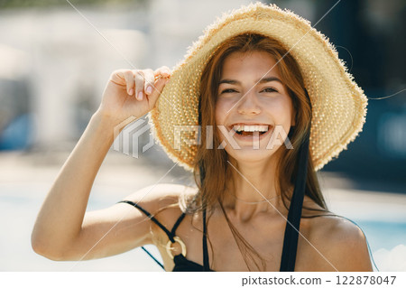 Young girl in a summer hat standing near swimming pool. Girl wearing black swimwear and a hat. Girl posing for a photo near the pool. 122878047