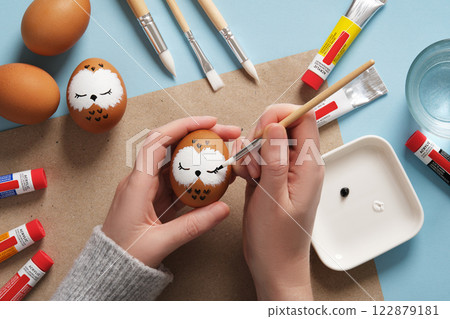 Drawing a sleeping owl on an eggshell. Step 2: Draw eyes. Preparation for Easter celebration. Acrylic paints in tubes, water, brushes. 122879181