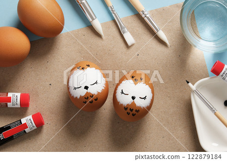 Easter eggs with sleeping owls, hand-painted with acrylics, on a background of paints, brushes and palettes. DIY concept Easter eggs with sleeping owls, hand-painted with acrylics, on a background of paints, brushes and palettes. DIY concept 122879184