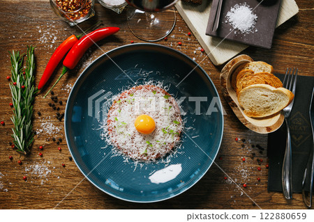 Exquisite beef tartare featuring a raw egg yolk and Parmesan cheese. Served with toasted bread, chili peppers, and rosemary. A perfect choice for a high-end dining experience 122880659