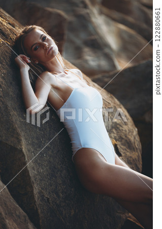 Sensual blonde woman in a white swimsuit lounging on rocks, exuding effortless confidence and beauty. 122880661