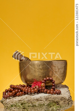 Copper singing bowl, wooden stick and prayer beads on stone, yellow background Copper singing bowl, wooden stick and prayer beads on stone, yellow background 122881473